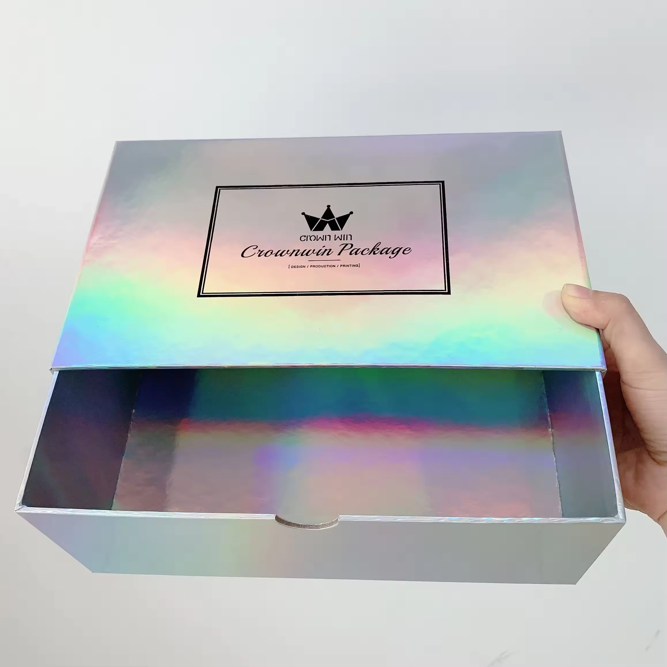A hand holding an open holographic foil box with a glossy surface displaying a rainbow effect. The box features a black label that reads “Crownwin Package”.