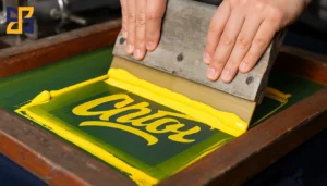 A person using a squeegee to spread yellow ink on a silk screen during a screen printing process. The screen has a green base with the word "Chato" partially visible in yellow cursive.